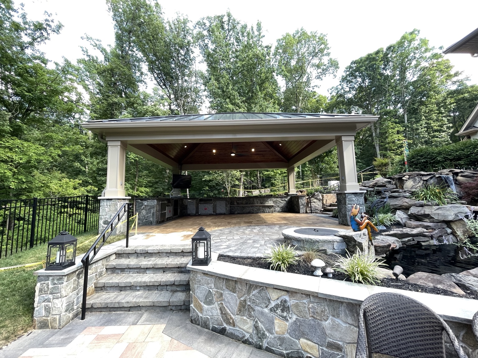 Covered outdoor pavilion with a stone kitchen, fire pit, paver patio, and a stacked-stone waterfall surrounded by trees