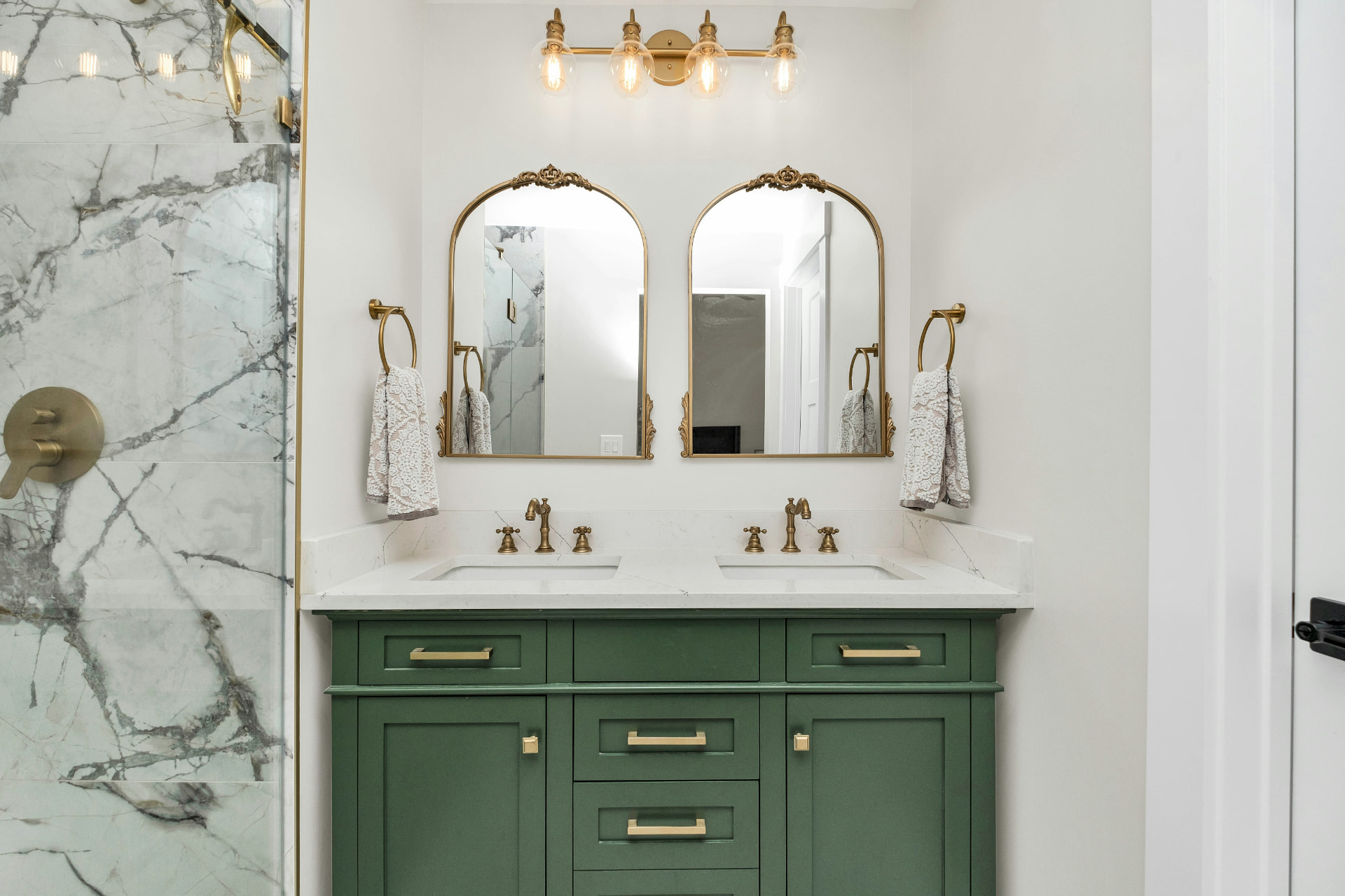 Bathroom with a forest-green double vanity, white countertop, arched mirrors, and brass fixtures beside a marble-tiled shower