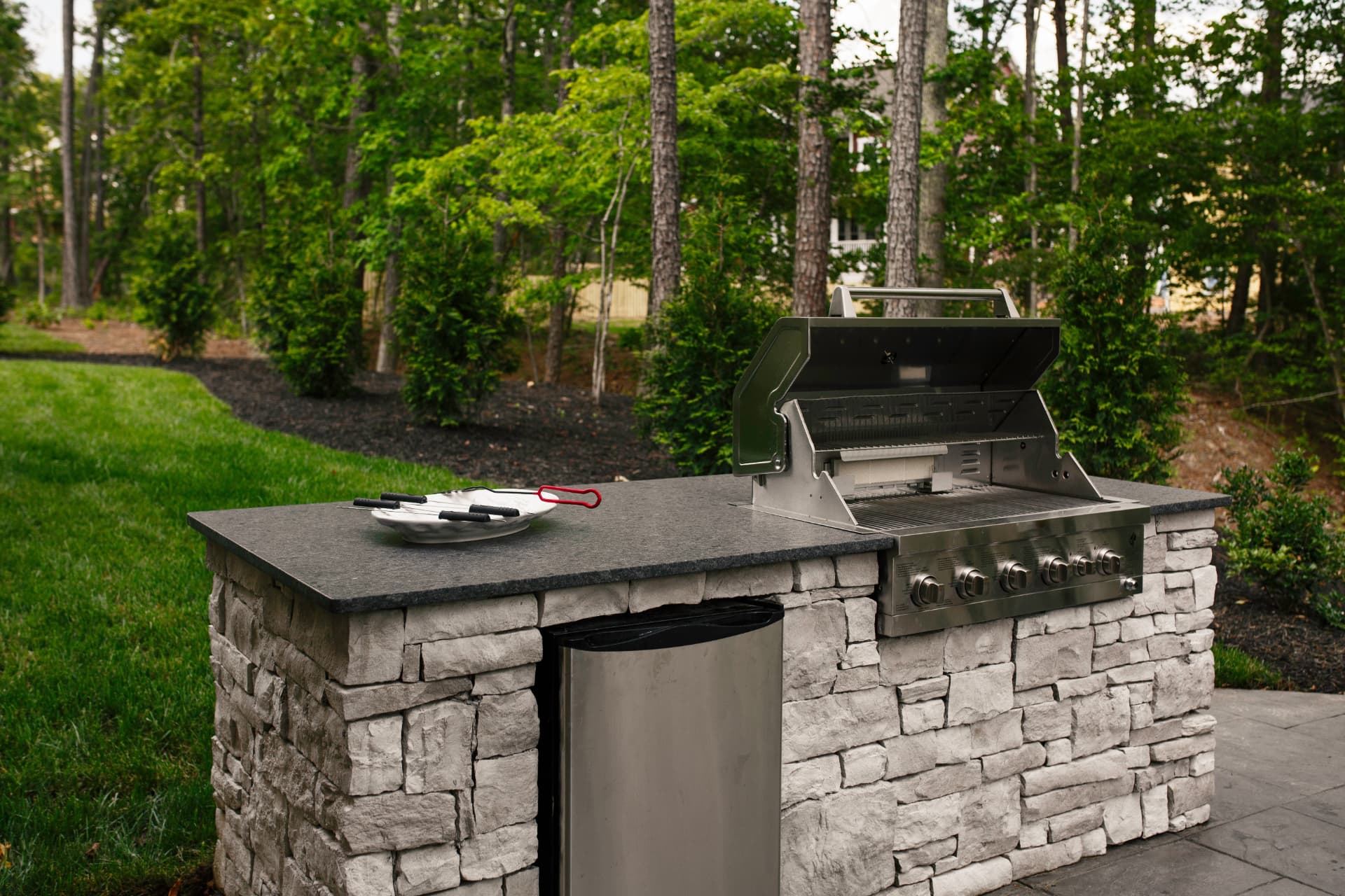 Stone outdoor kitchen with a built-in stainless steel grill, dark countertops, and a wooded backyard