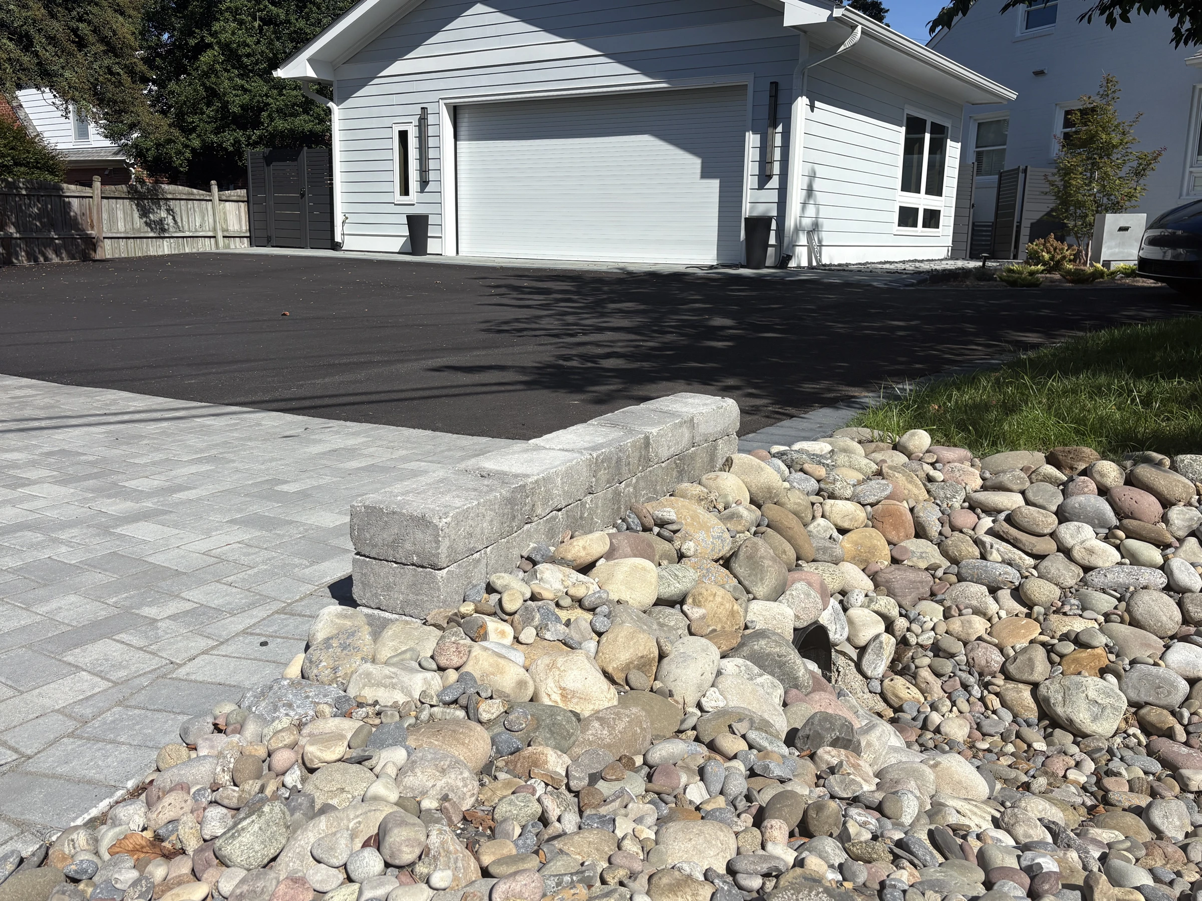 Gray paver apron meeting an asphalt driveway beside a low block wall and river-rock drainage bed