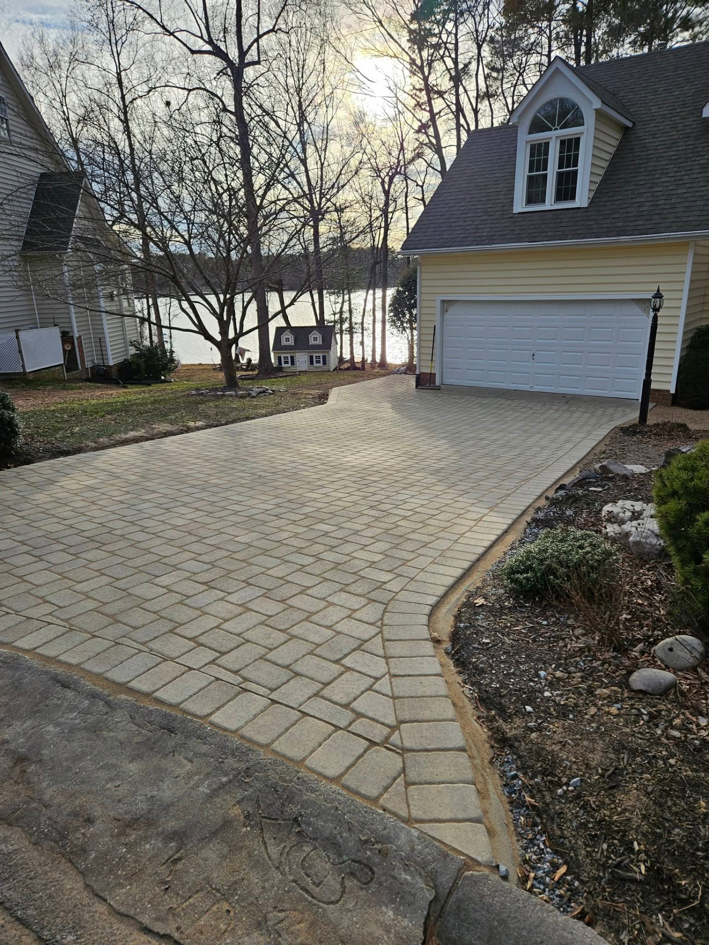 Tan paver driveway curving past garden beds toward a lakeside garage
