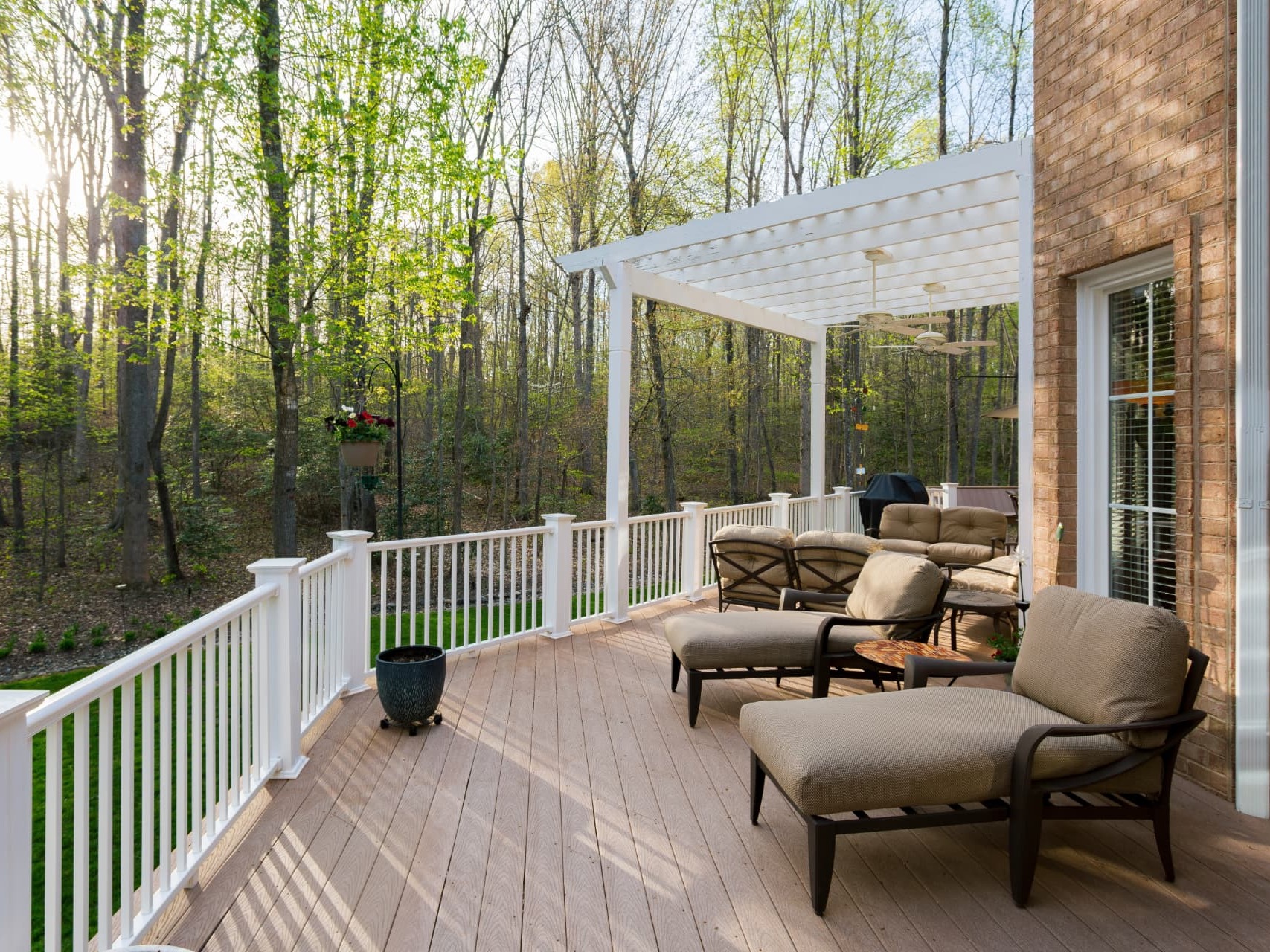 Covered wood deck with a white pergola, comfortable seating, and a wooded backyard