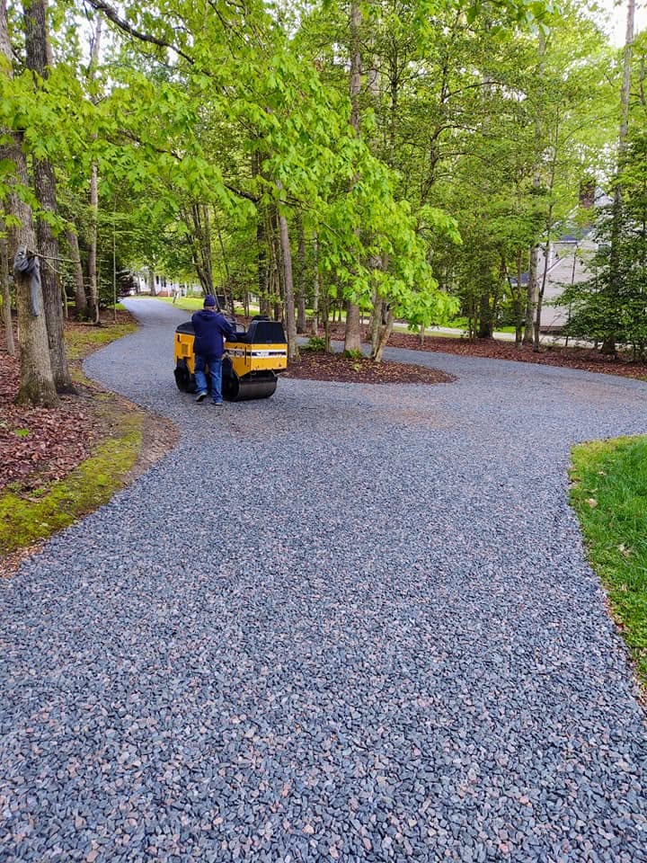 Gravel driveway base being compacted with a roller beneath a canopy of spring trees