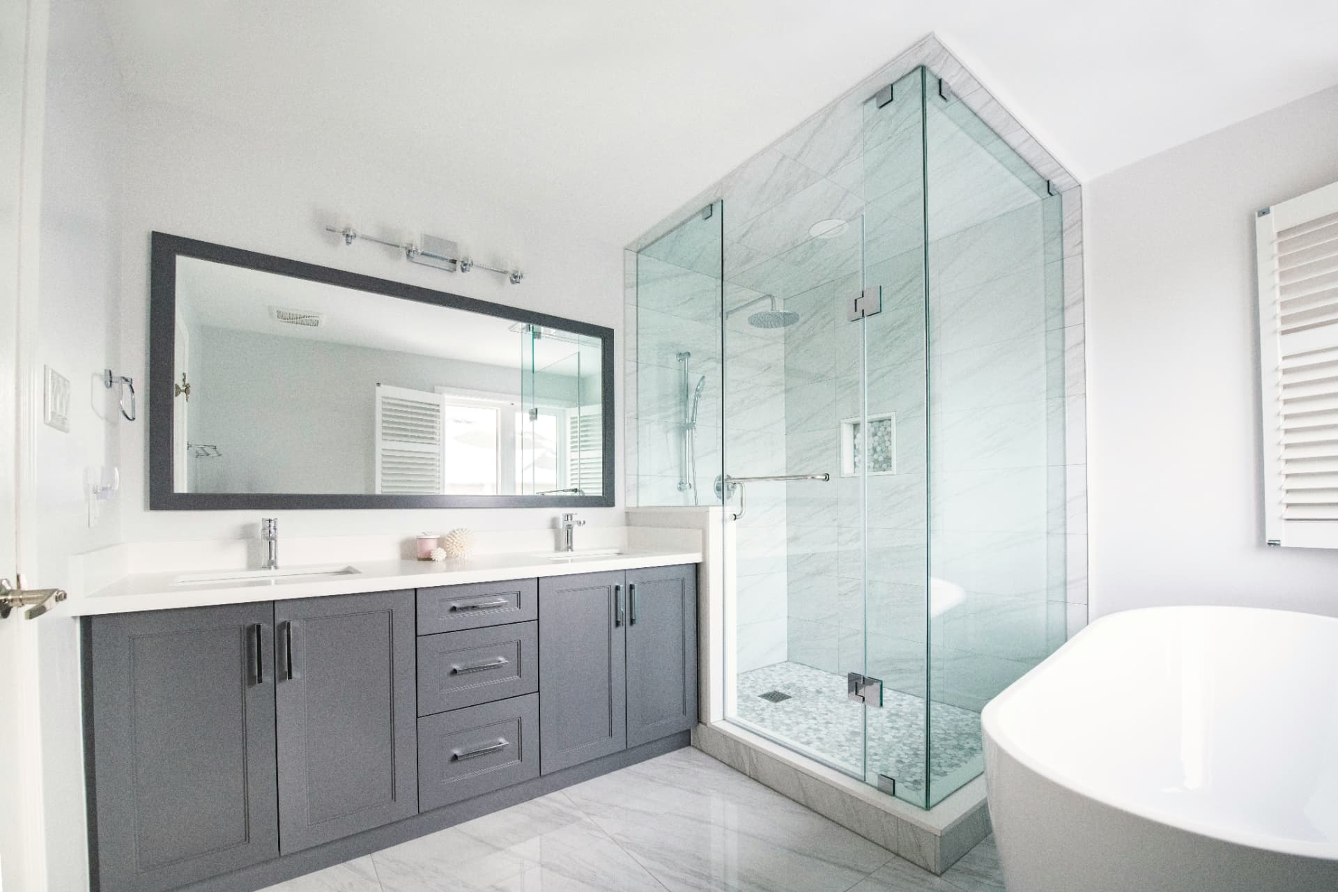 Bathroom with a charcoal double vanity, a glass corner shower, and the edge of a freestanding tub beside a window with shutters