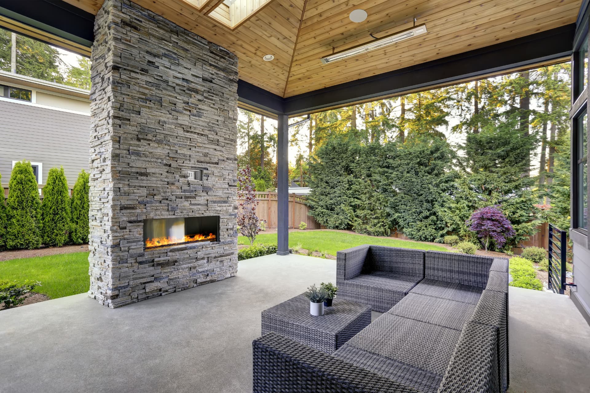 Covered outdoor living room with a floor-to-ceiling stacked-stone fireplace, warm wood ceiling, woven lounge seating, and a green lawn framed by mature trees