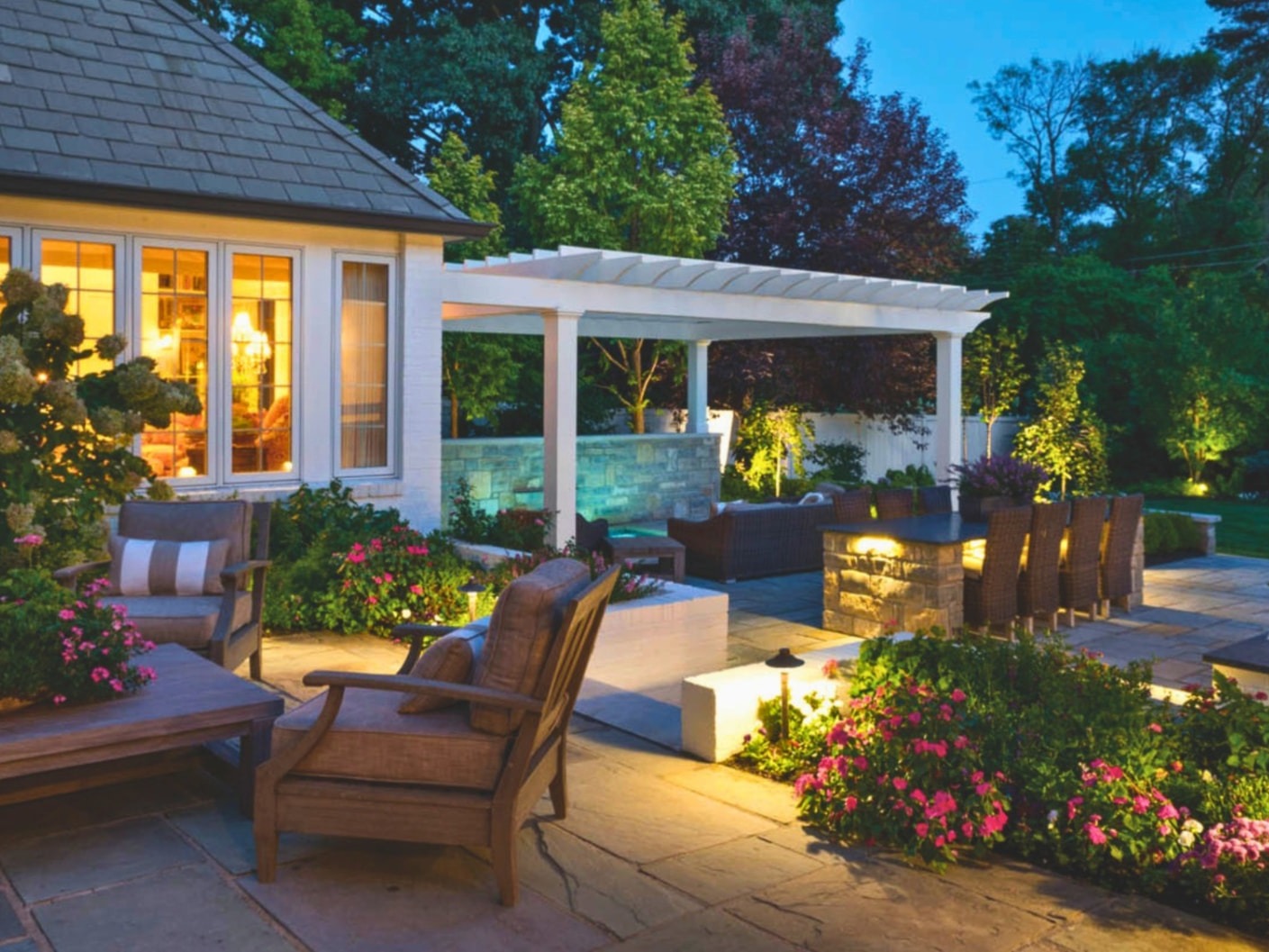 Evening patio beside a home with glowing windows, a white pergola, stone fire feature, outdoor dining area, and warm landscape lighting