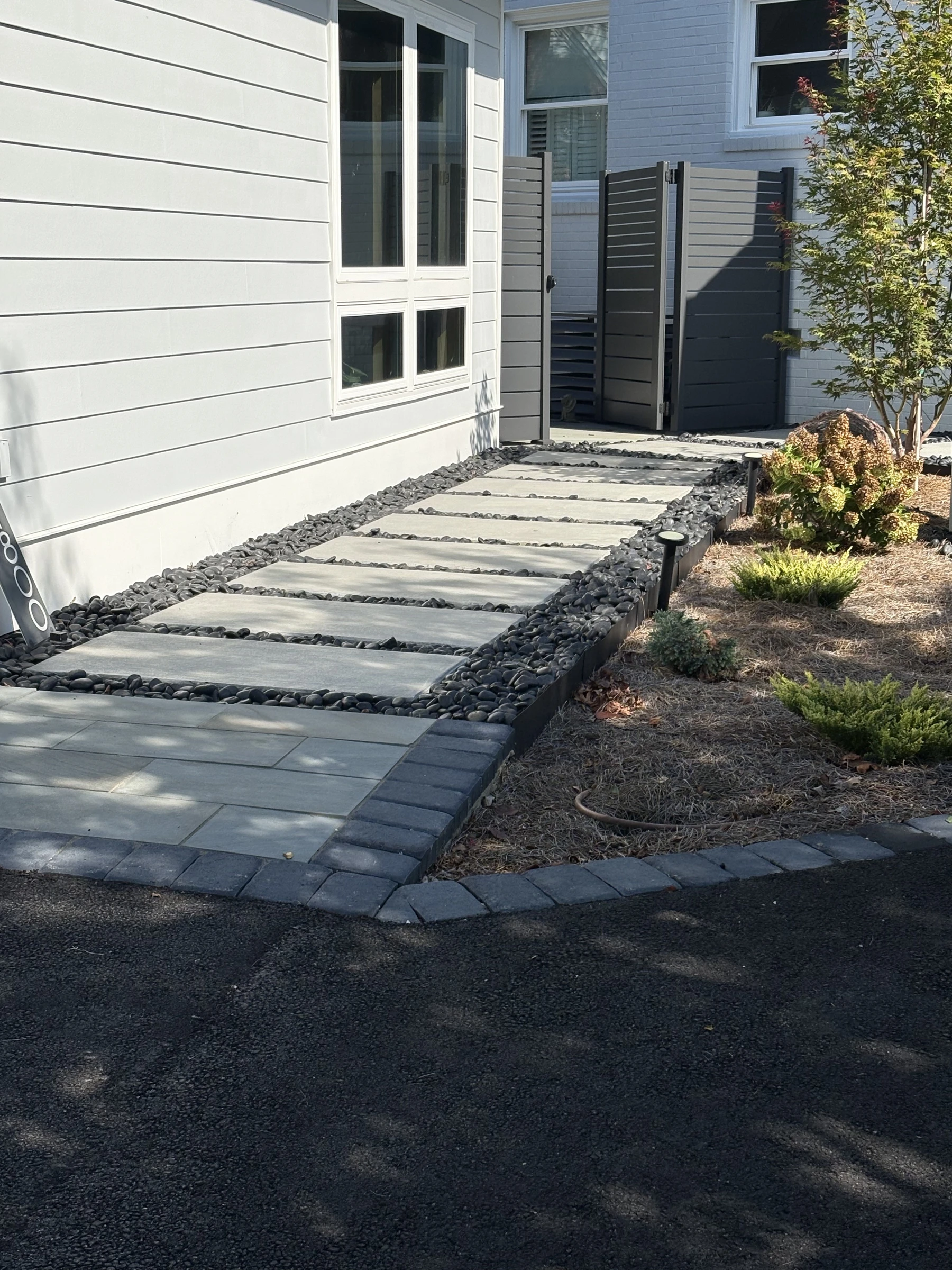 Concrete slab walkway set in dark stone with a paver border connecting the asphalt driveway to a gray gate