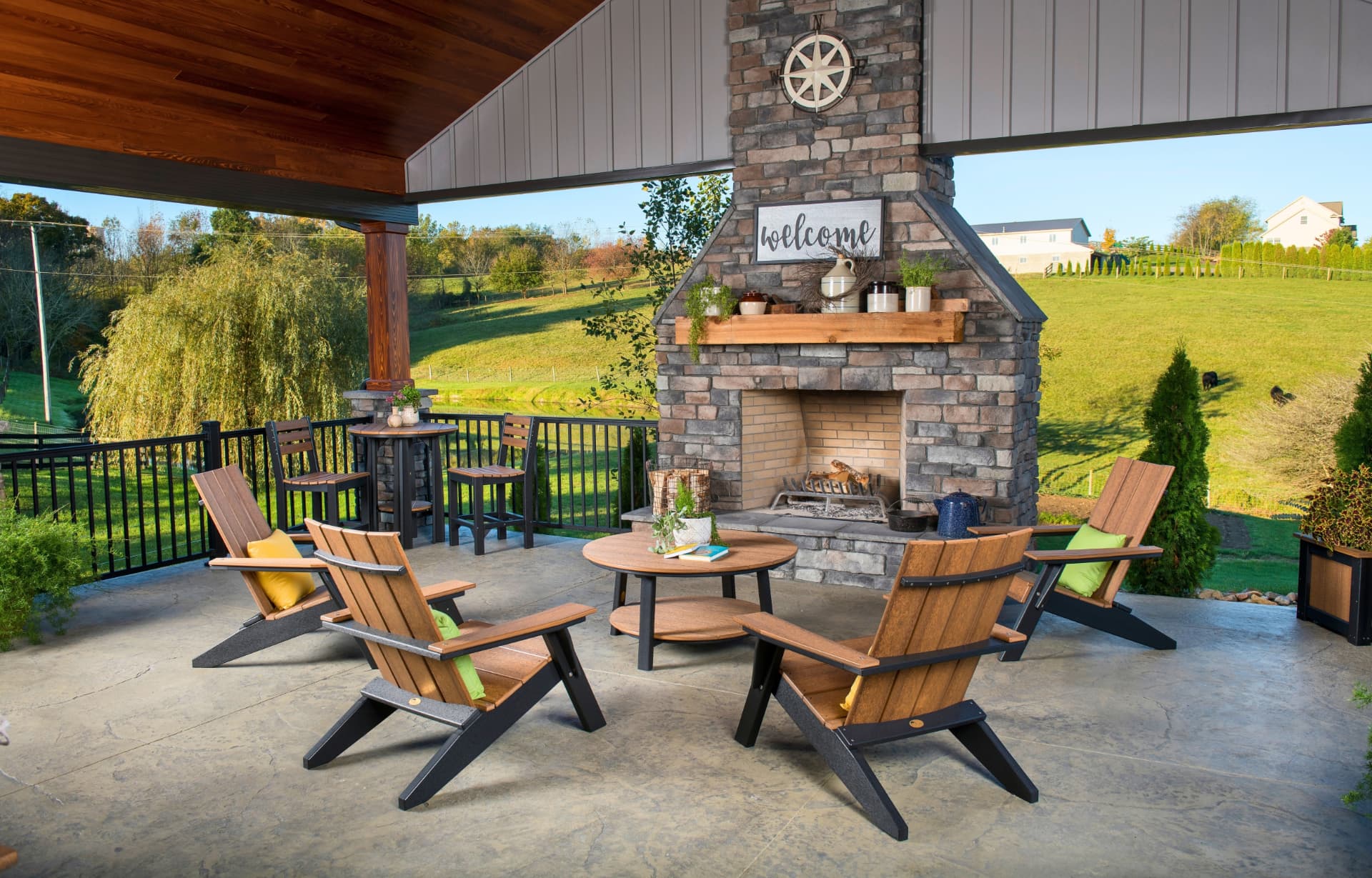 Covered patio with a stone fireplace, wooden Adirondack chairs, and rolling green pasture in the background