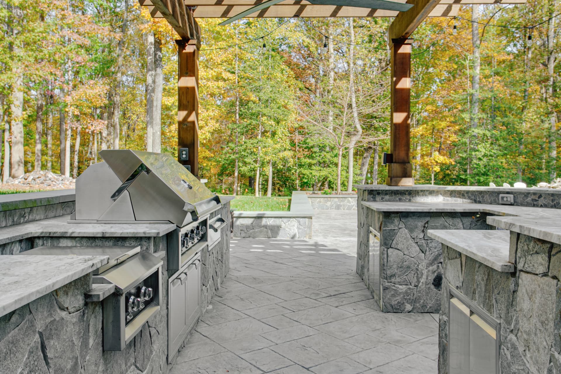 Covered outdoor kitchen with stone counters, stainless steel grill, and a wood pergola overlooking a paver patio