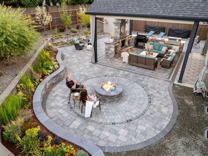 Overhead view of a paved outdoor living space centered on a circular fire pit with seating, surrounded by a pergola, outdoor seating, and soft landscaping